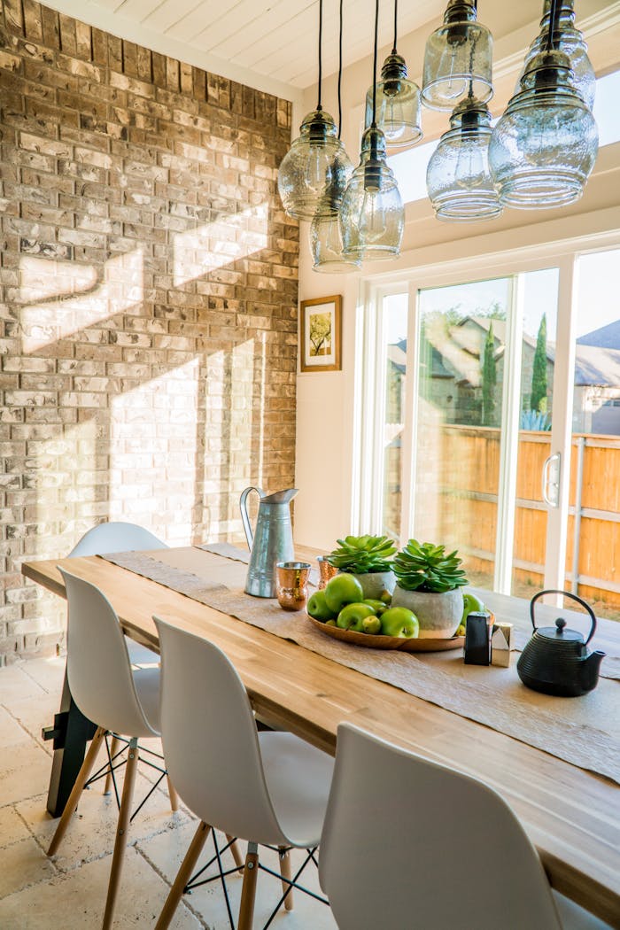 Sunlit dining room with contemporary decor and a rustic brick wall, featuring stylish lighting and a wooden table.